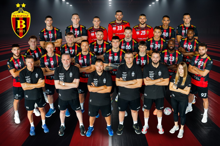 A group of athletes and staff pose together in a handball team photo, wearing matching jerseys and black training gear on a blue court.